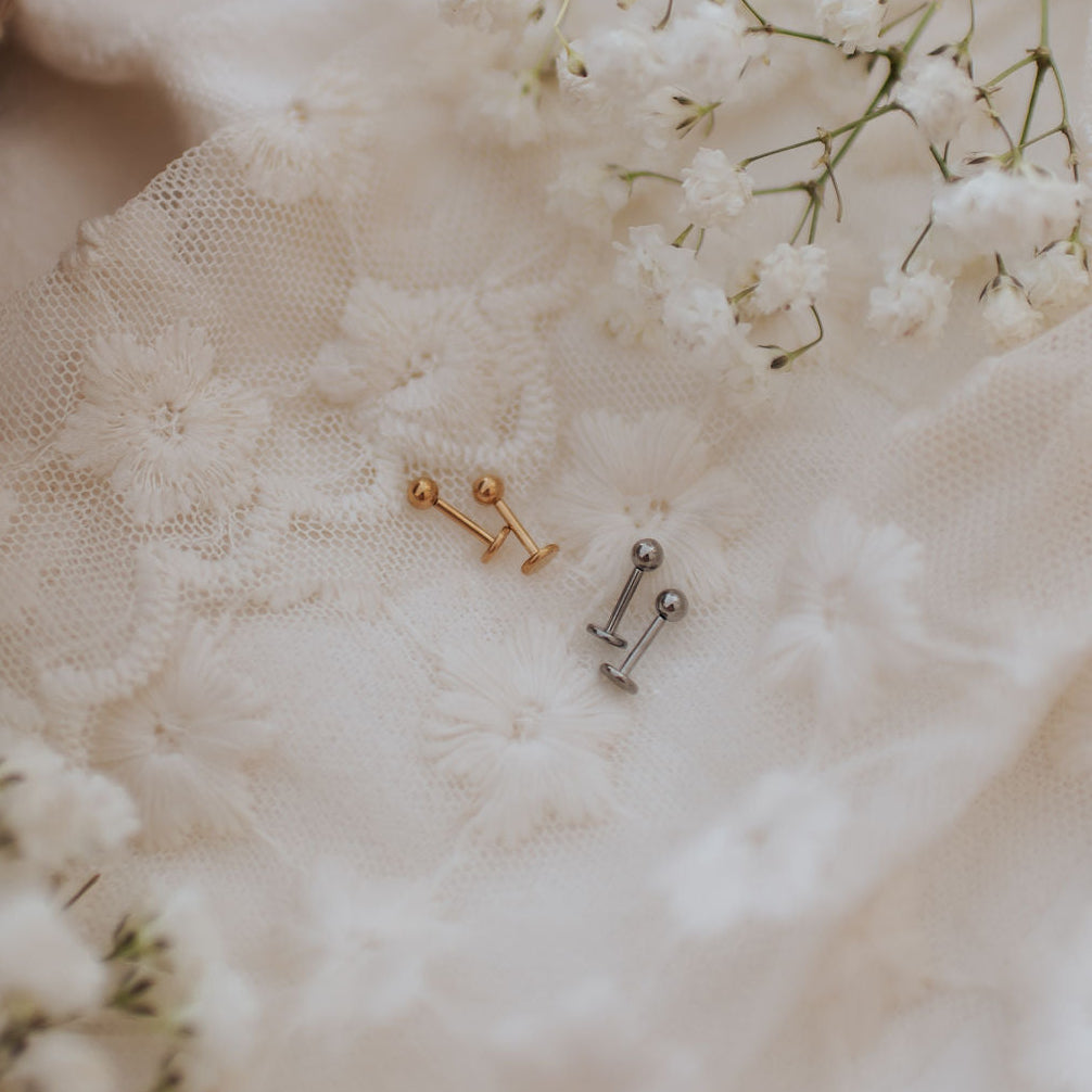 Two small barbell stud earrings, one gold-toned and one silver-toned, displayed on textured white lace fabric with white baby's breath flowers.