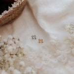 Two small stud earrings, one silver-toned and one gold-toned, displayed on a white lace fabric with baby's breath flowers and a woven basket of red fruit in the background.