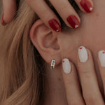 Close-up of a person's ear with earrings, hand touching hair, and various nail colors.