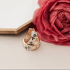 Two gold tea cup inspired earrings with gemstones on a white surface next to a pink rose.