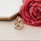Two gold tea cup inspired earrings with gemstones on a white surface next to a pink rose.