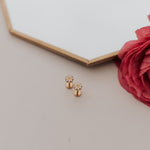 Gold daisy earrings on a light surface with a red flower and hexagonal mirror in the background