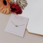 Gold necklace with a blue stone on a Mauve Jewelry Co. card with flowers in the background