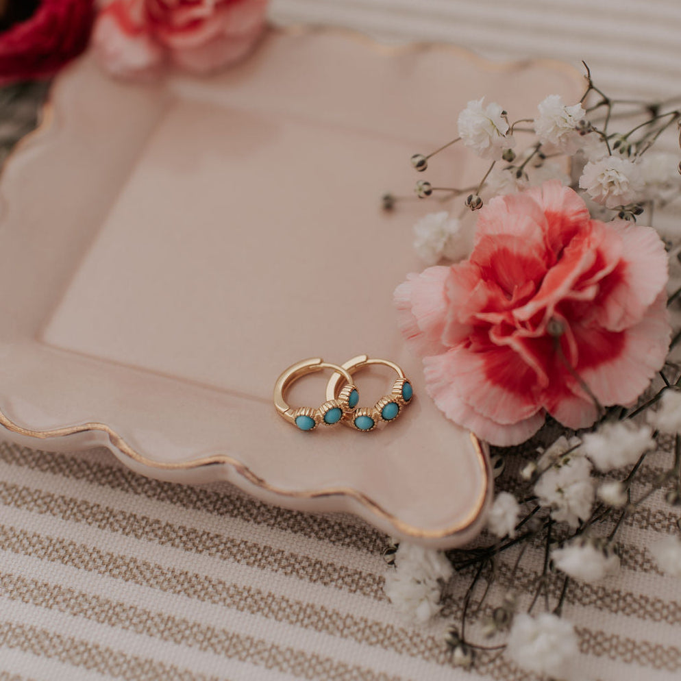 Two gold earrings with turquoise stones on a decorative plate with flowers and baby's breath.