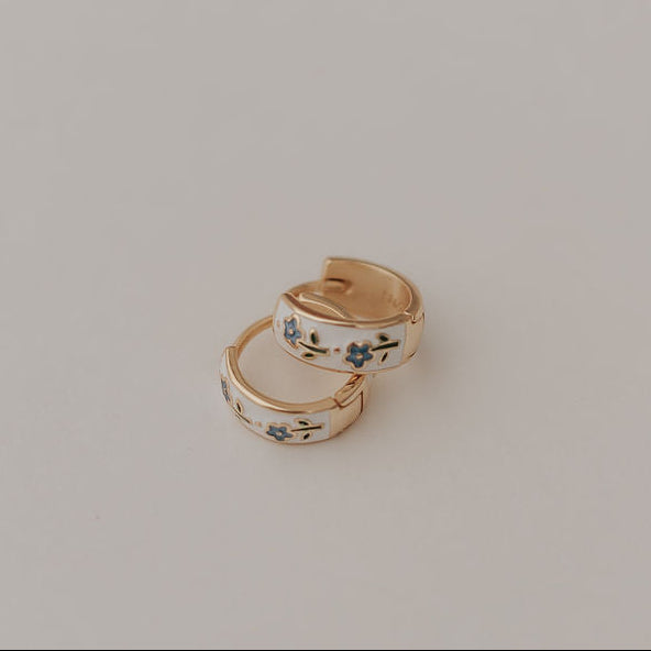 Two gold earrings with blue floral designs on a light beige background