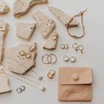 Gold jewelry including rings, earrings, and necklaces on stone and concrete surfaces with a Mauve Jewellery Co. pouch.