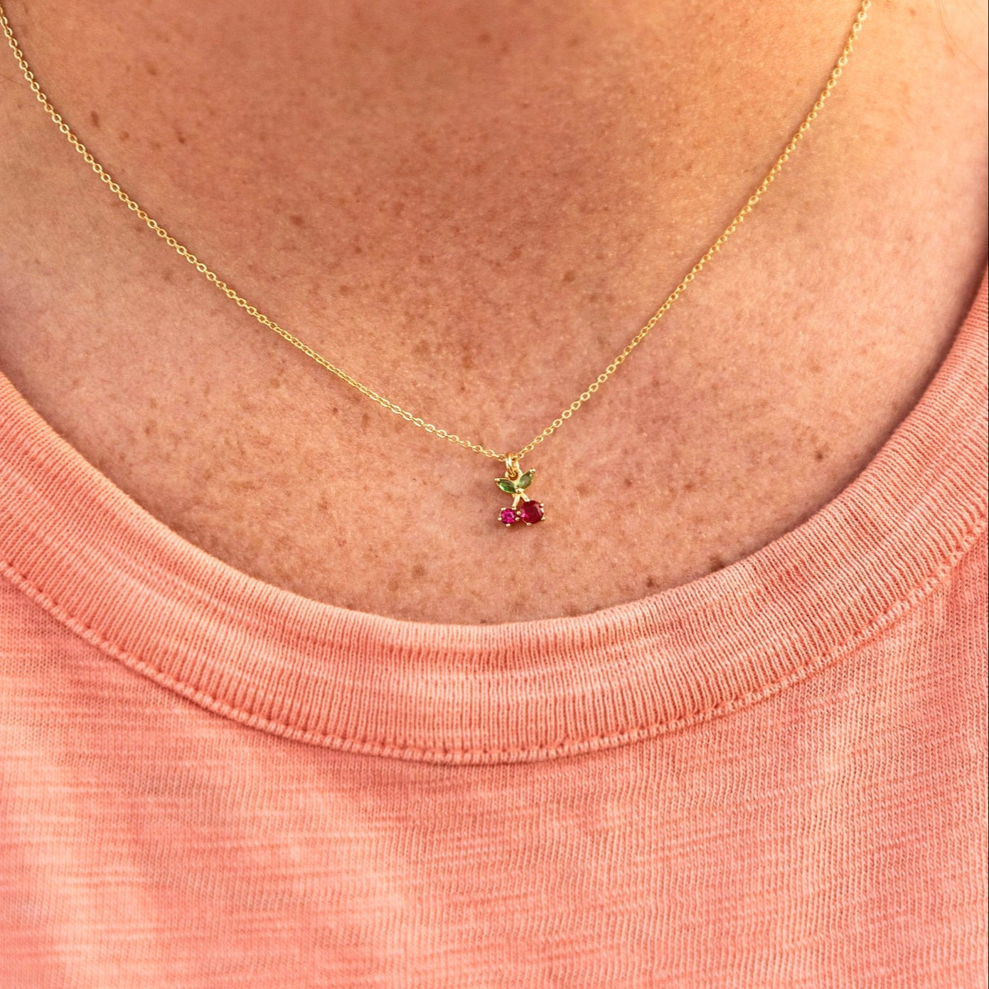 Necklace with a small red cherry pendant worn by a person in a pink shirt.