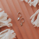 Small hoop earrings with cross charms on a textured pink surface with white tassels.