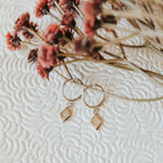 Gold hoop earrings with diamond charms on a textured white surface with dried flowers.